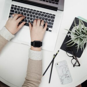 person typing on laptop on table next to potted plant, glasses, two pencils, and an iPhone
